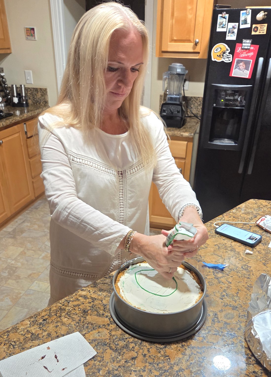 Meredith standing at her kitchen counter, squeezing a tube of green gel icing onto a cheesecake, the first outline just begun.